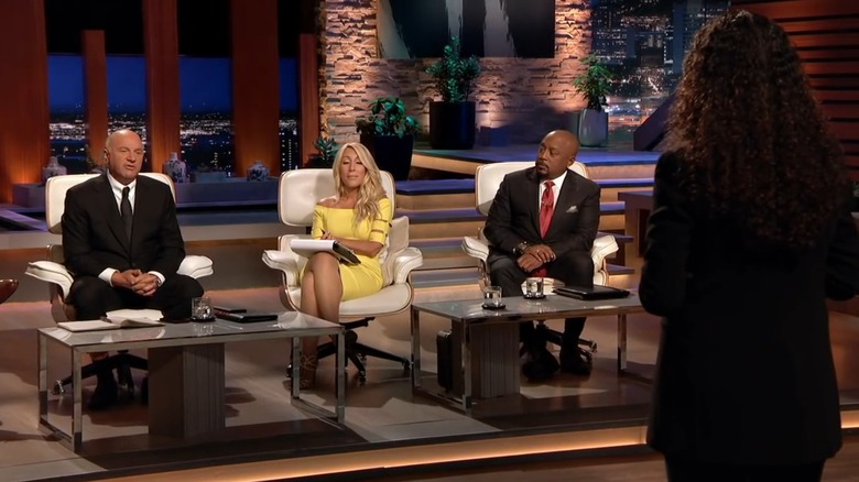 Shark Tank investors Kevin O'Leary, Lori Greiner, and Daymond John sitting on the set during a pitch. Professional lighting in the Shark Tank television studio. Focus on venture capital negotiation and high-equity deals. Representing the iconic reality TV moment where the Goalsetter pitch took place. Financial literacy app founder presenting to celebrity investors. The tension of startup funding and valuation on national television. Famous Sharks evaluating a fintech business model. Historic context for the app's growth beyond reality TV. Shark Tank set with prominent celebrity investors during a season 10 pitch session. Capital investment and entrepreneurship television program context for Goalsetter.