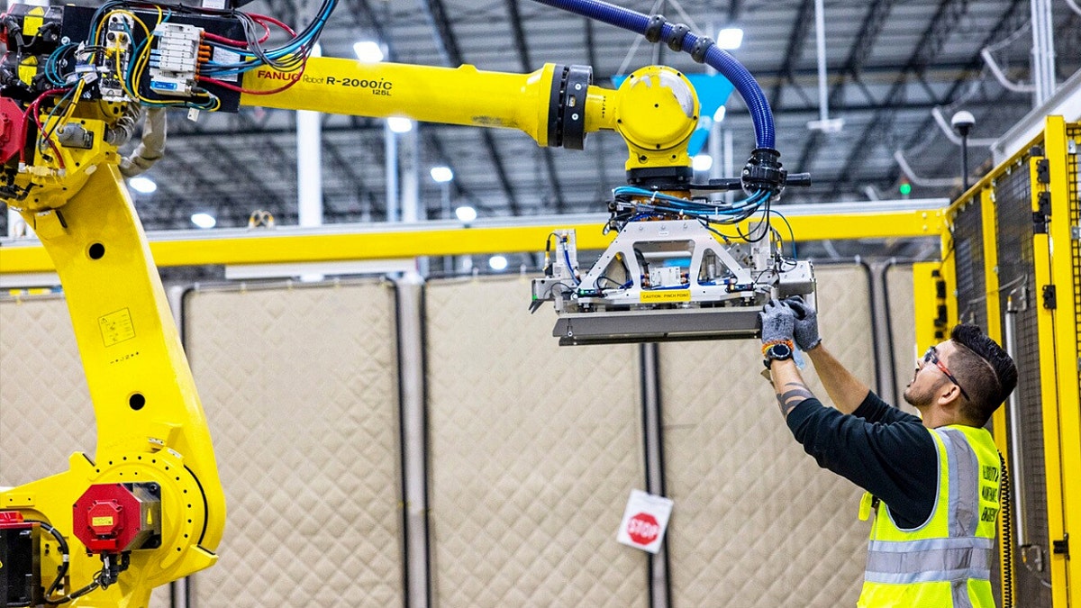 An Amazon warehouse worker inspecting a Blue Jay robot unit on the floor.