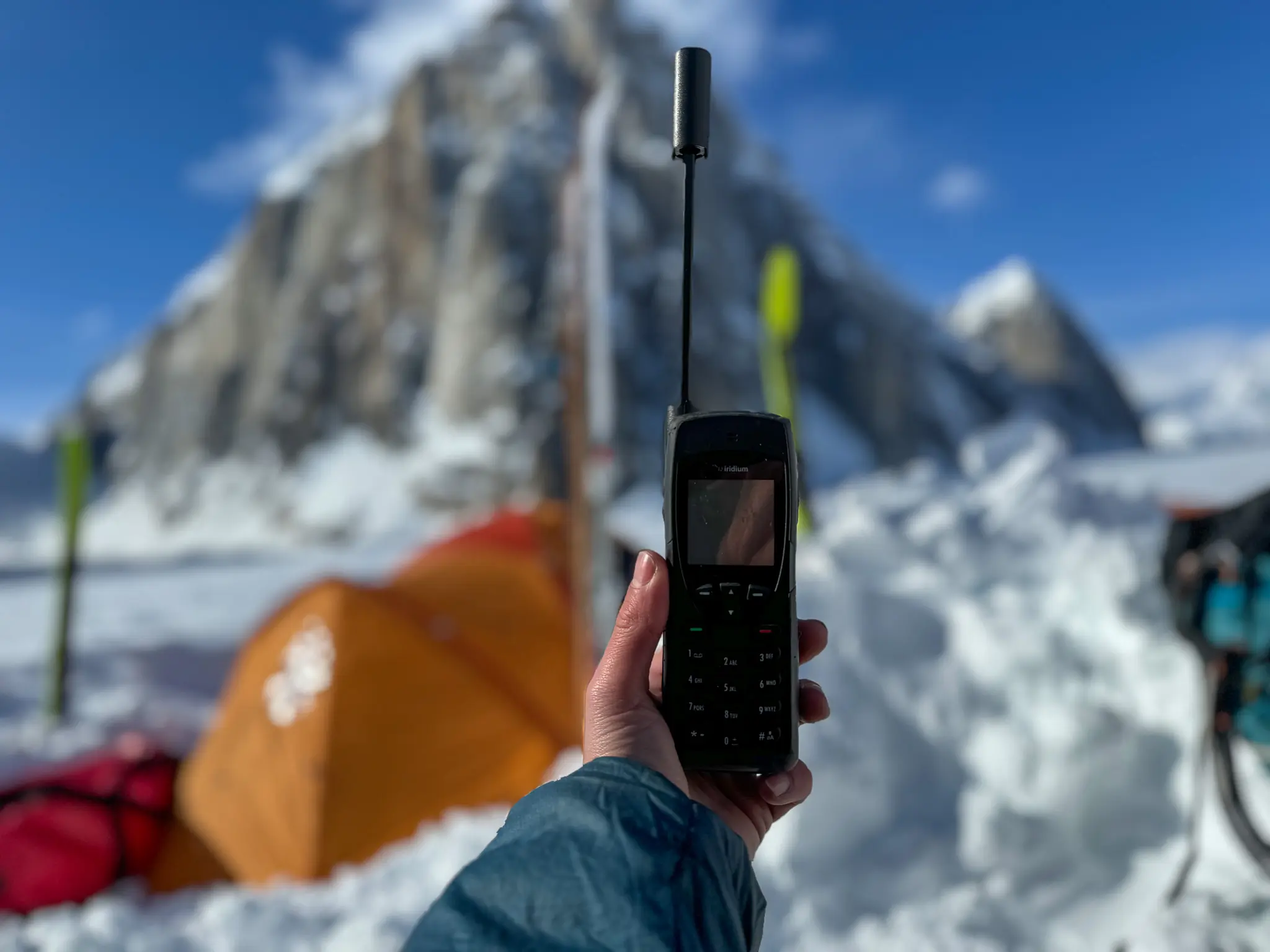 A person holding an Iridium satellite phone against a background of rugged, snow-capped mountains.