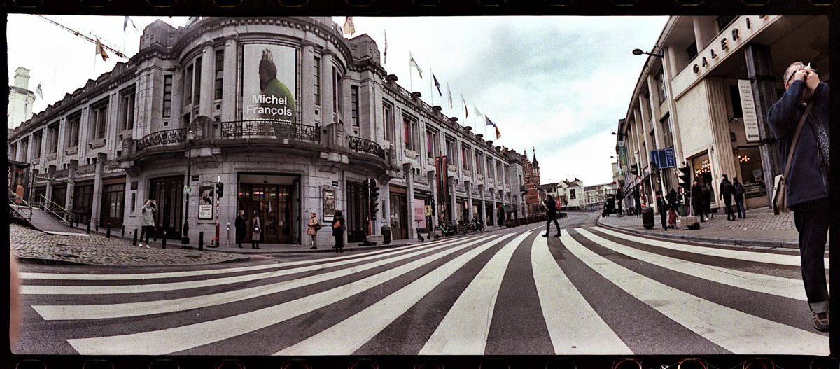 A panoramic photo of a street crossing showing curved horizontal lines and a blurry finger in the corner.
