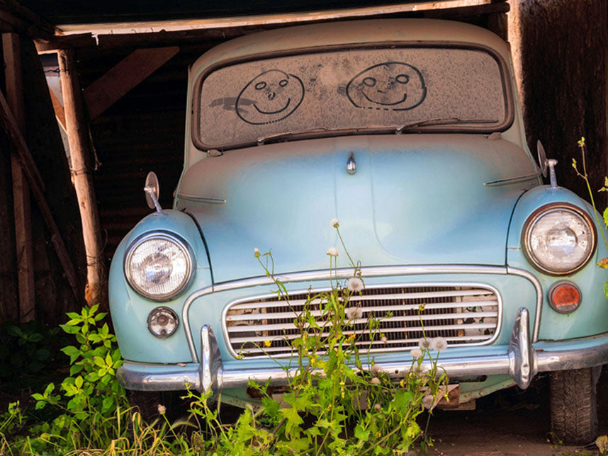 An old classic car covered in thick layers of dust and debris inside a dark storage building.