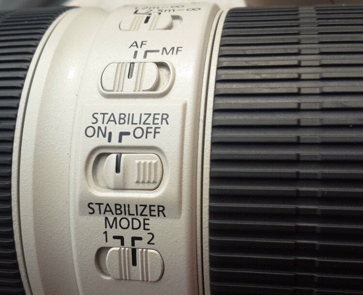 Close-up of a camera lens with the Image Stabilizer (IS) switch being moved to the 'Off' position.