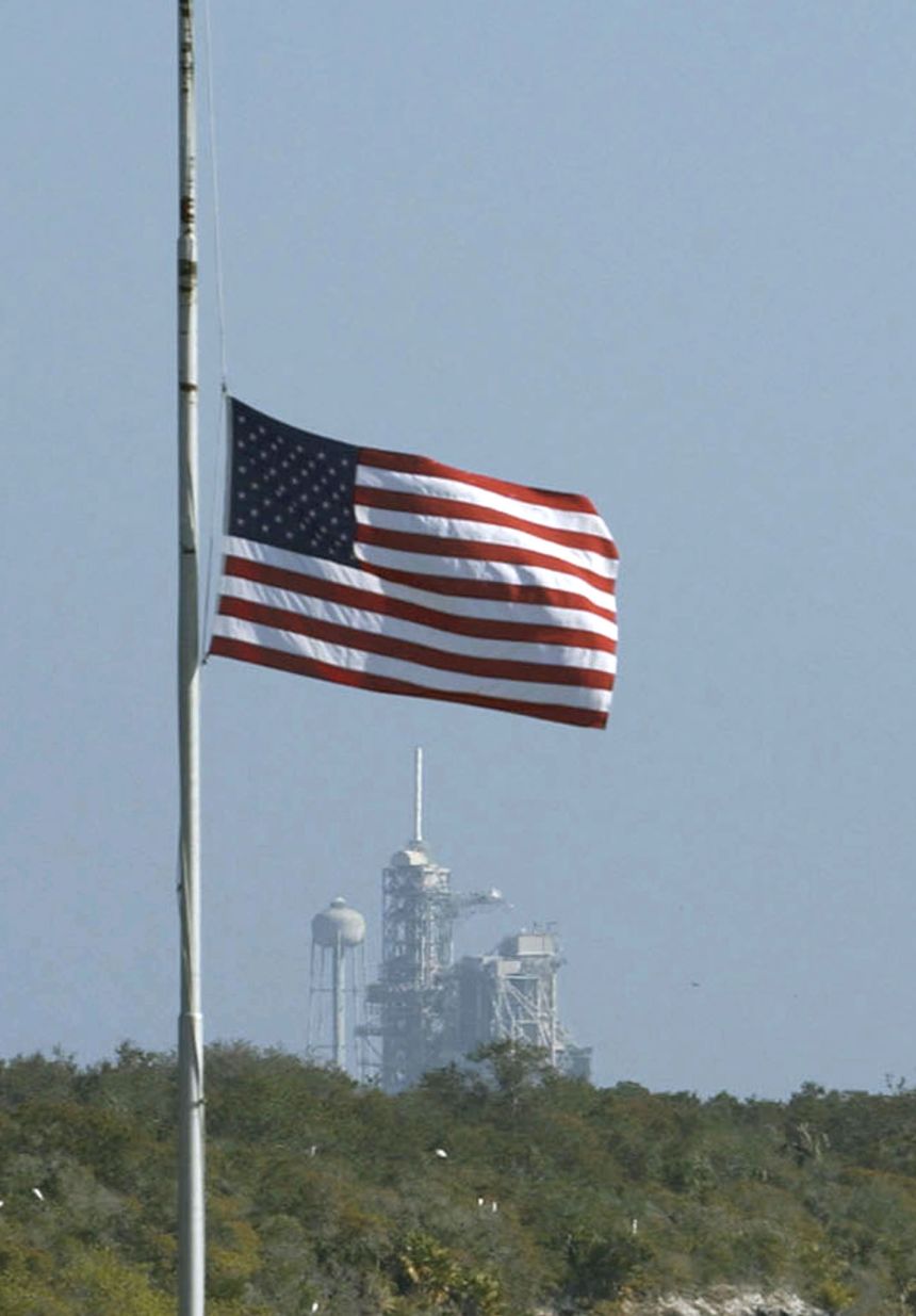 The American flag flying at half-staff at the Kennedy Space Center with the Vehicle Assembly Building in the background.