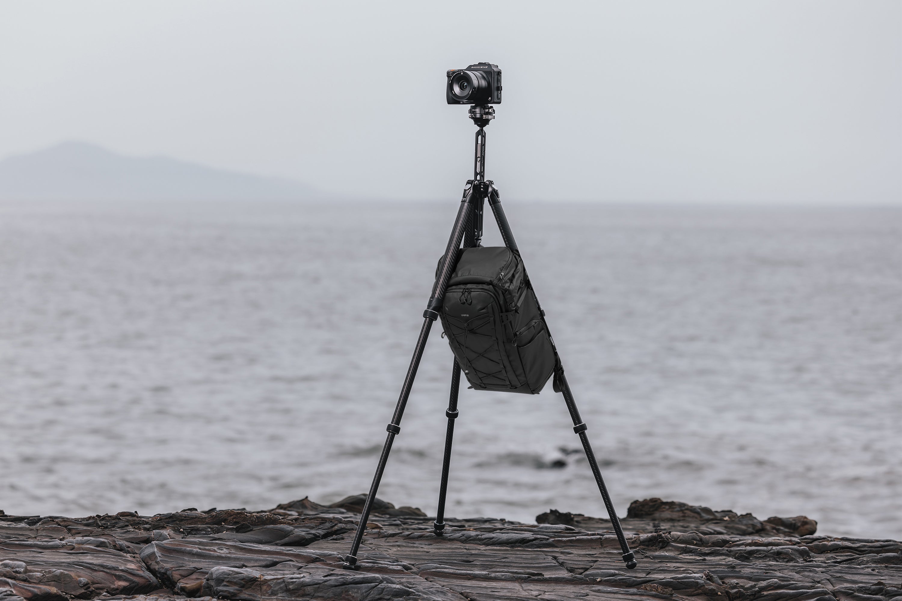 A camera tripod with a black gear bag hanging from its center column hook in an outdoor setting.