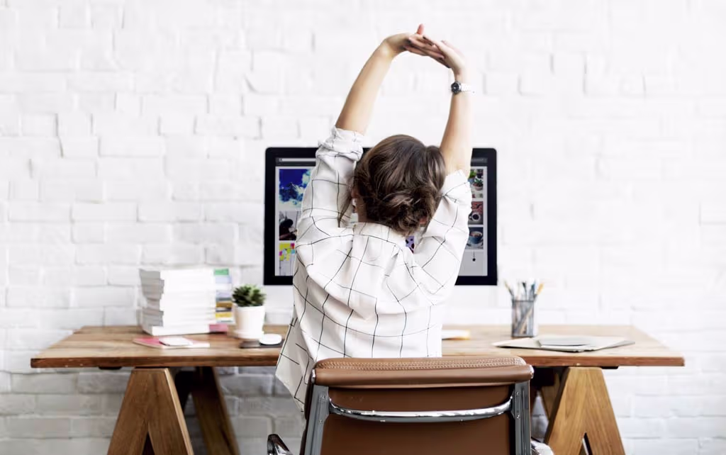 An office worker standing and performing overhead stretches in a brightly lit office.